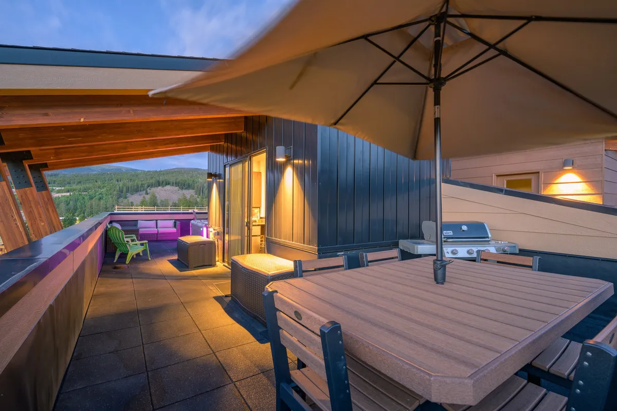 A spacious deck with an outdoor dining table, grill, inviting green chairs, umbrella, and a hot tub—set against a backdrop of open sky. The arrangement invites both al fresco dining and relaxation, with a seamless transition between eating, socializing, and unwinding.