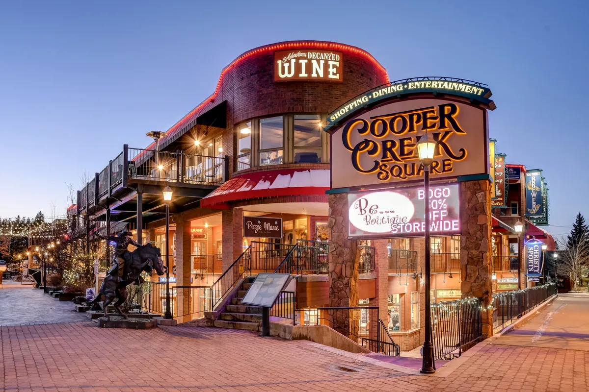 This exterior image captures the lively atmosphere of Cooper Creek Square, featuring a prominent sign, wine shop, outdoor seating areas, and a distinctive horse sculpture. The scene suggests a bustling hub of dining, shopping, and entertainment under festive decor.