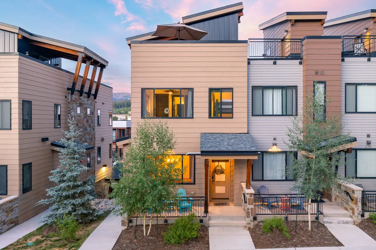 The image features the modern exterior of a single-family penthouse villa in Winter Park, Colorado, showcasing sleek architecture, a beautifully landscaped front yard, and a spacious outdoor seating area. The porch is set up for relaxing and enjoying sunset views, with comfortable seating that invites guests to unwind or socialize with neighbors. The inviting scene hints at memorable evenings spent hosting barbecues and sharing stories against a breathtaking mountain backdrop.