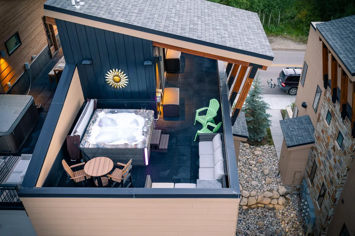 A tranquil rooftop space featuring a hot tub, stylish outdoor seating area with a green chair and wooden table, sunburst wall décor, and an open sky above. The ambiance hints at both relaxation and space for gathering with friends beneath the stars.