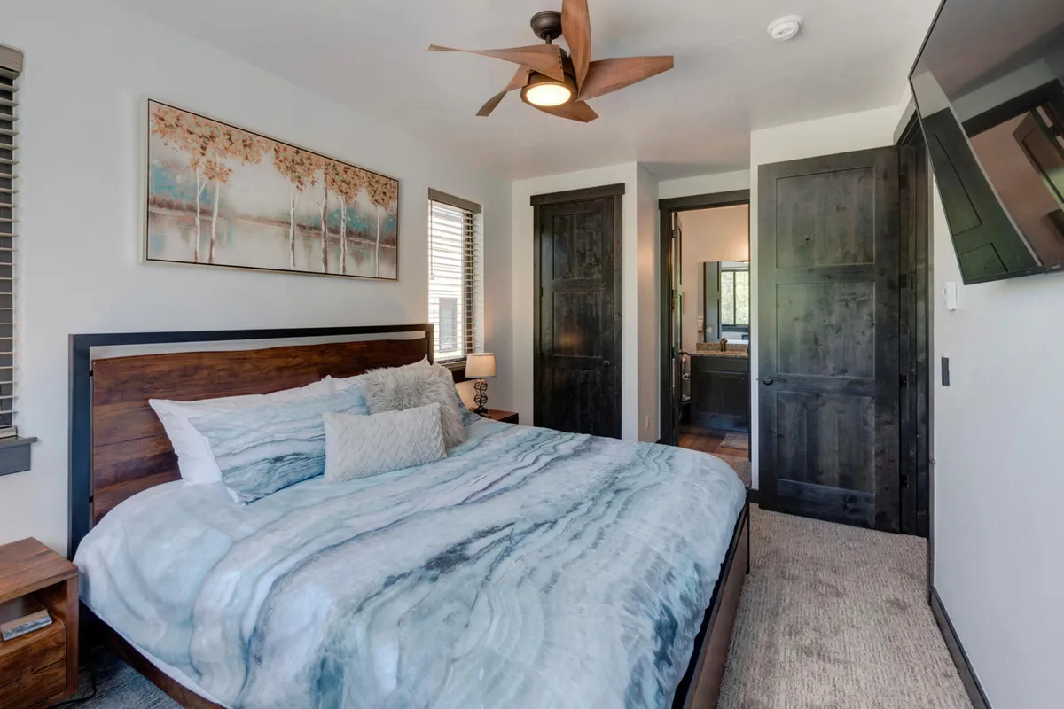 A serene bedroom featuring a bed with a marbled comforter, artwork above the bed, a ceiling fan, and a wall-mounted TV. Soft natural light fills the room, creating a tranquil and cozy retreat for relaxation or entertainment.