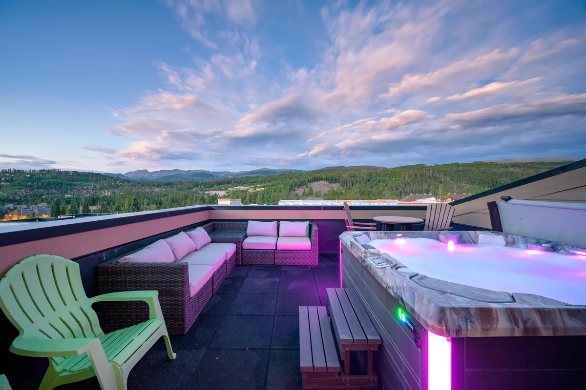 A welcoming rooftop deck featuring a modern hot tub with warm, inviting lighting, surrounded by cozy patio chairs and a stunning view of the mountains in the distance. The scene is set for both relaxing soaks and social gatherings, with the sun setting over the peaks.
