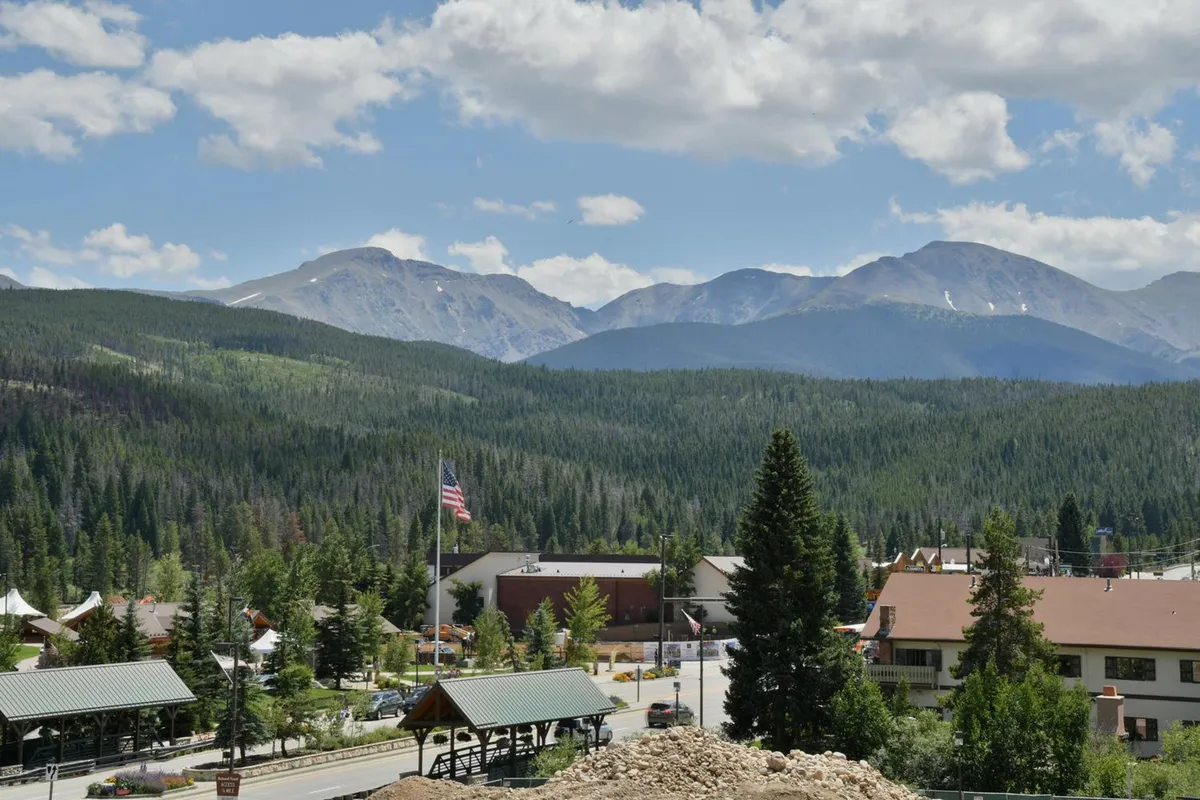 A panoramic view capturing majestic mountains, lush forests, and a small town nestled in the valley. A flag waves gently, hinting at local festivities and the spirit of exploration. The image evokes a sense of discovery and connection with nature.