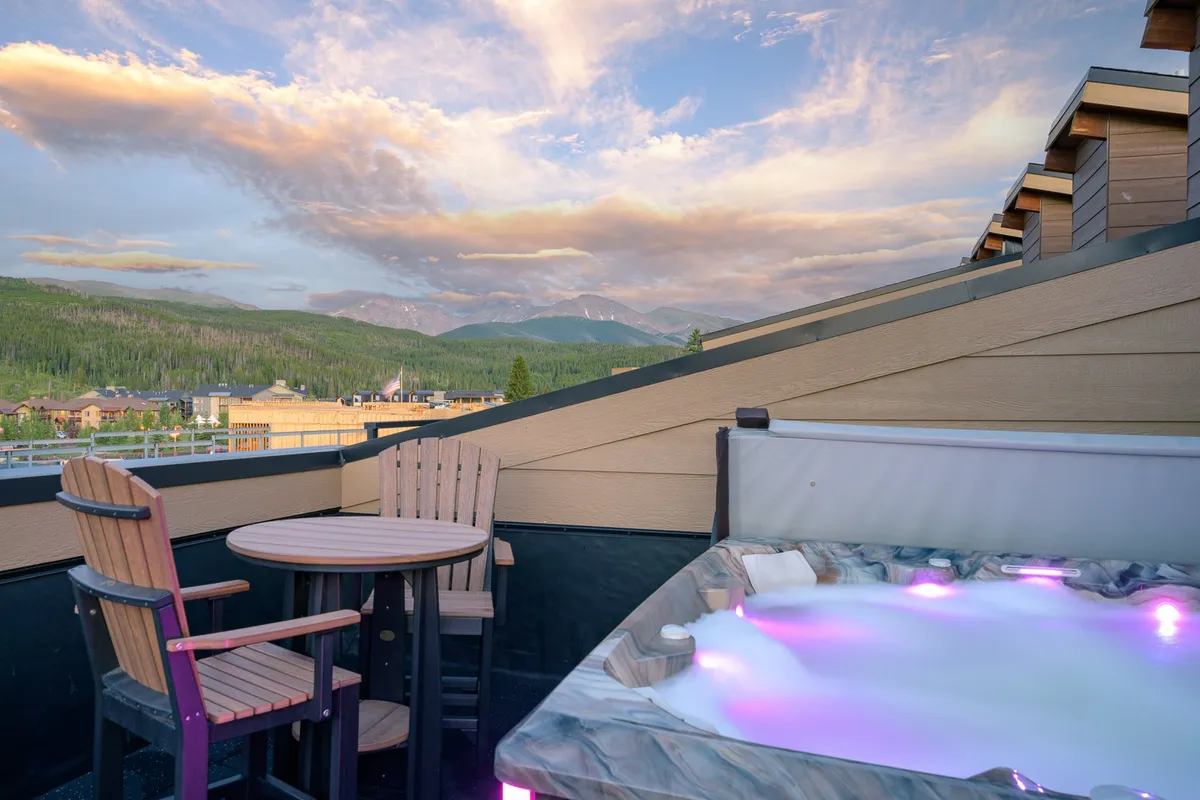 This image captures a rooftop jacuzzi glowing with inviting lights, surrounded by wooden chairs and a table. The setting is framed by a stunning mountain backdrop and an expansive sky with drifting clouds, evoking a tranquil and luxurious atmosphere. The lighting suggests either sunset or early evening, emphasizing both relaxation and breathtaking views.