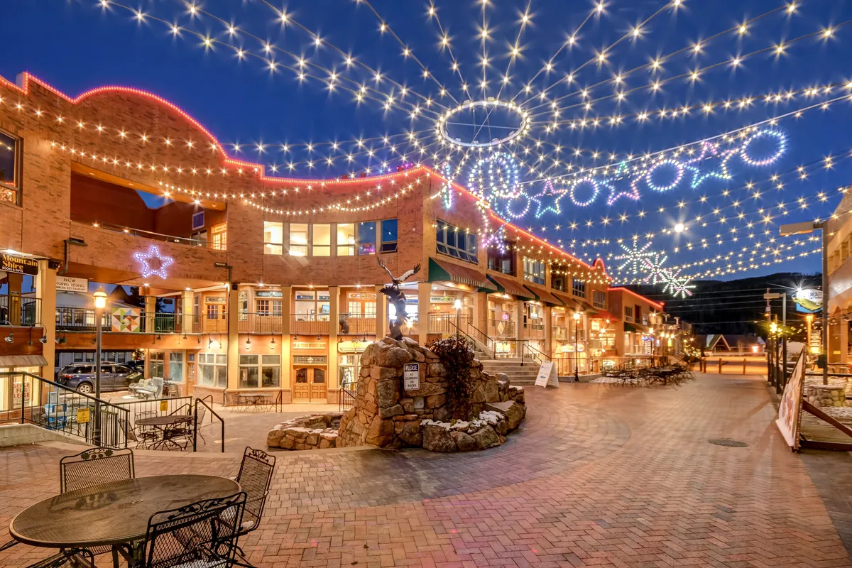 A charming scene of a brick pathway lined with string lights, decorative stars, and a central stone fountain, inviting visitors to stroll, dine, and gather with friends. The festive lighting conveys a sense of community celebration and lively downtown evenings.
