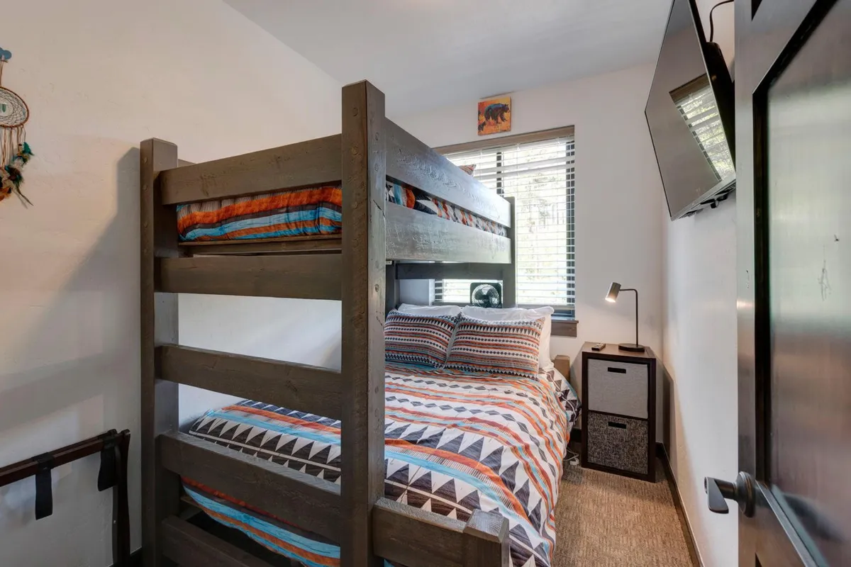 A playful bunk bed setup with colorful bedding, a TV, and a window with blinds. The space is bright and cheerful, designed for both relaxation and entertainment, ideal for kids or groups seeking memorable sleepovers.